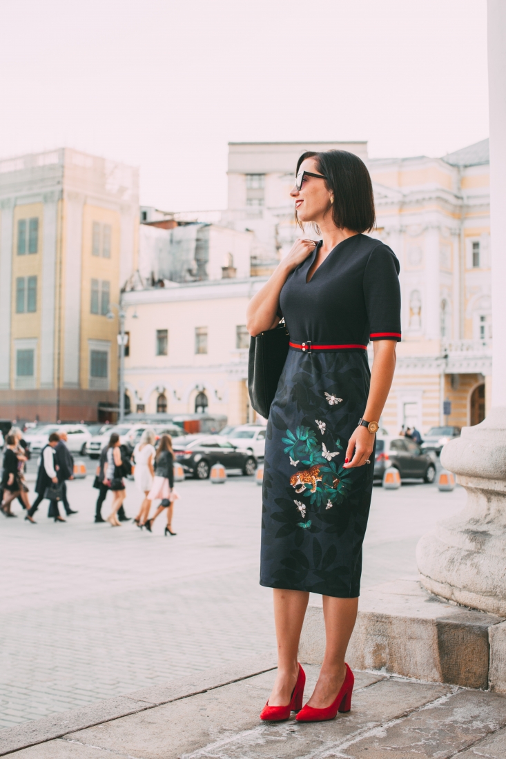 Photo shows a woman wearing a black dress with butterflies and a leopard on the skirt, with a black purse and red shoes