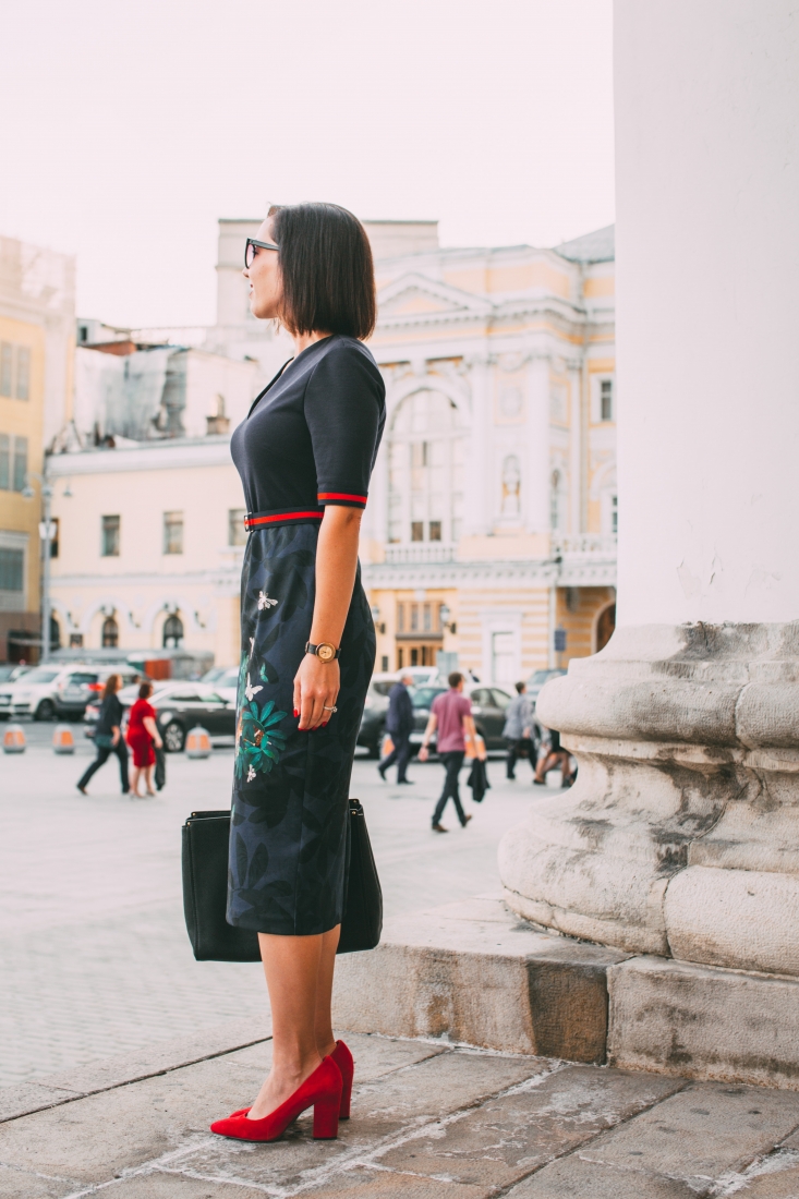 A woman poses on the street of a Moscow city square. She stands facing away from the camera, showing the side silhouette of slimming black dress with red accents and dressy red heels.