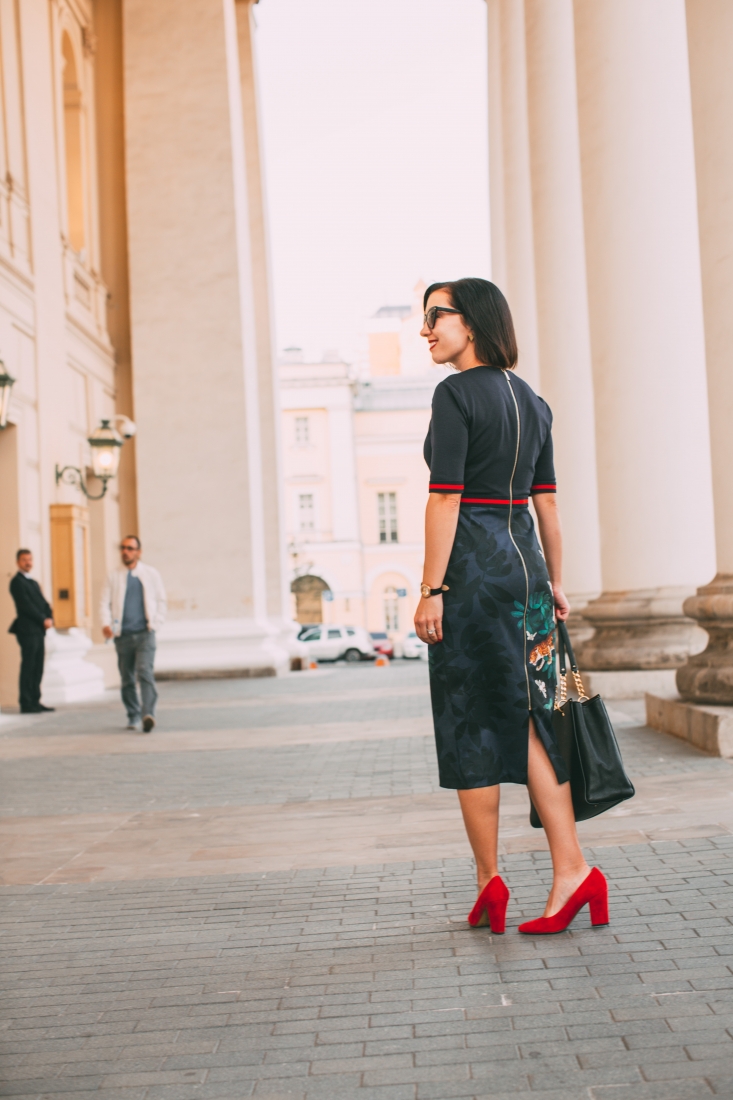 A woman poses with her back to the camera, walking along a city street. She's wearing a long black dress with a gold zipper detail along the back, paired with dressy red heels.