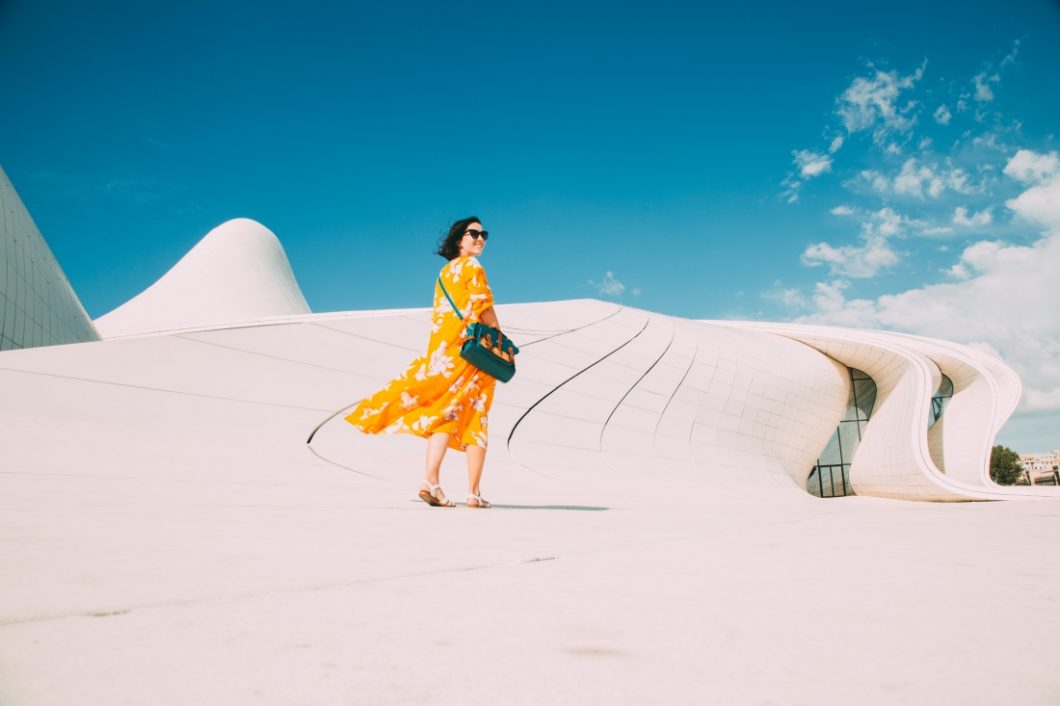 A woman wearing a bright marigold floral dress walks along a concrete pathway on a clear, sunny day in Baku, Azerbaijan.