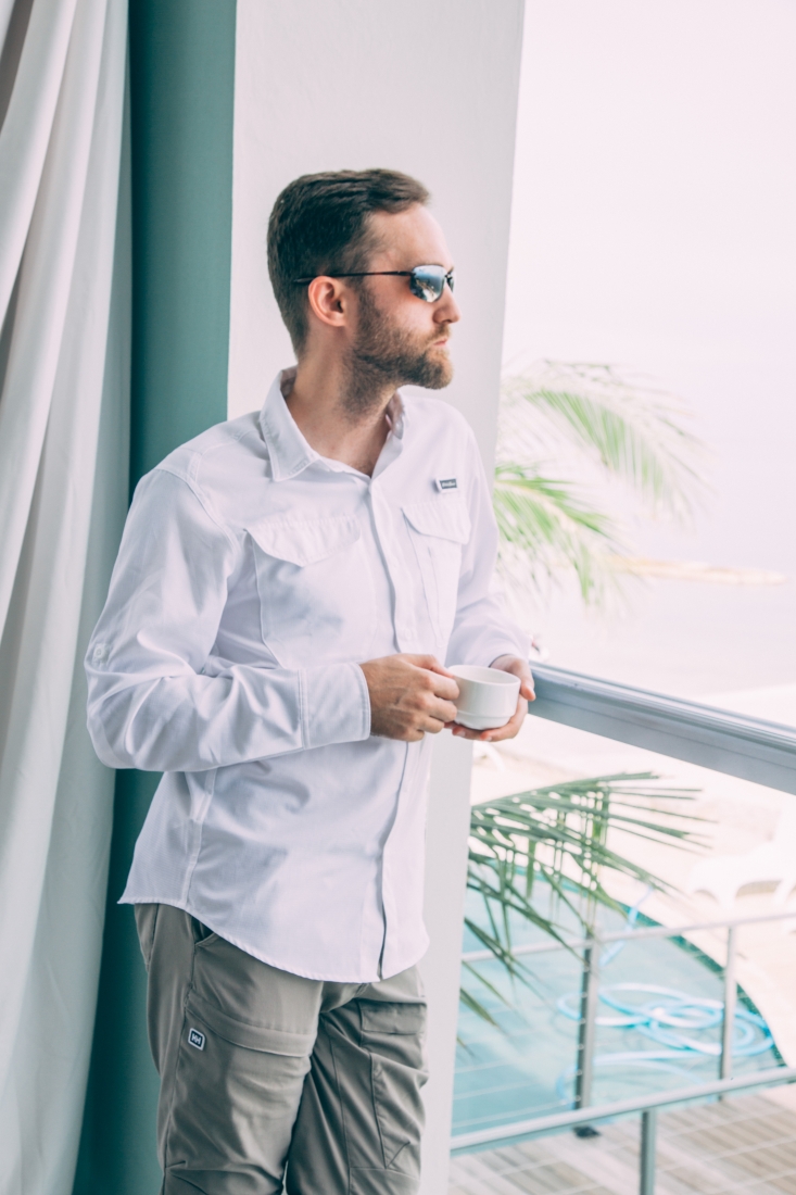 Zac wearing a white button down holding a coffee mug, while looking at the ocean