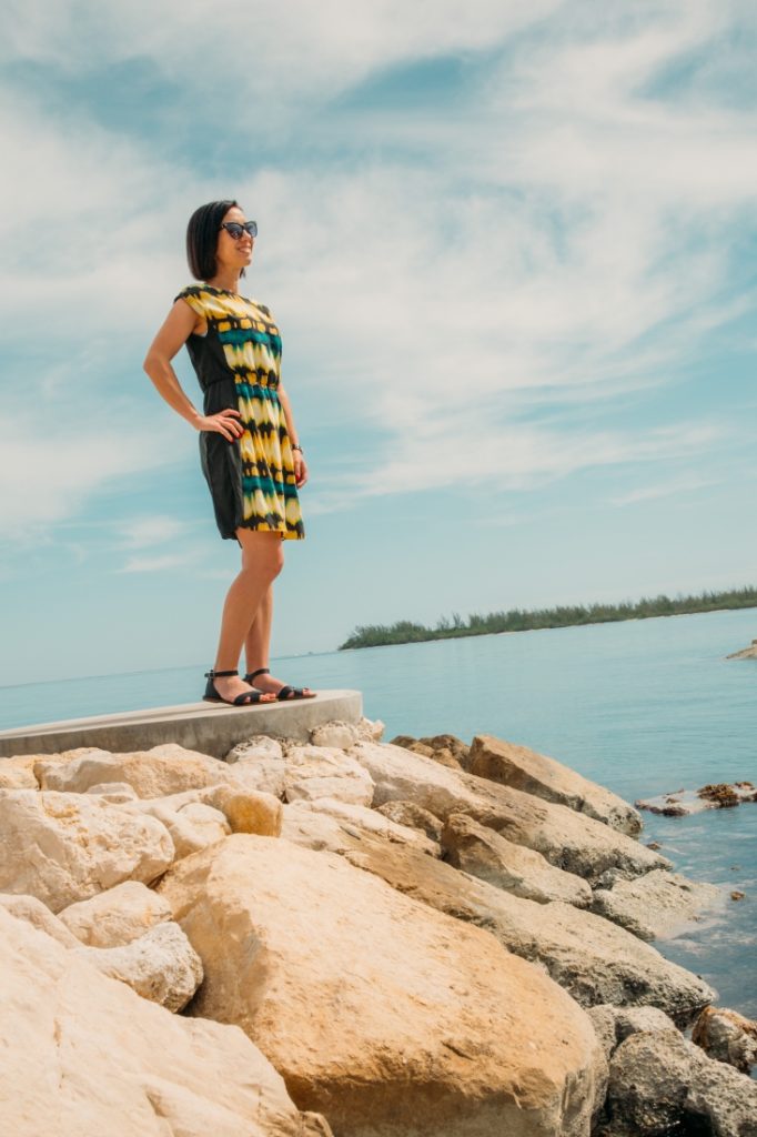 Lindsey wearing a colorful sundress and black sandals standing on a rock by the ocean on a sunny day