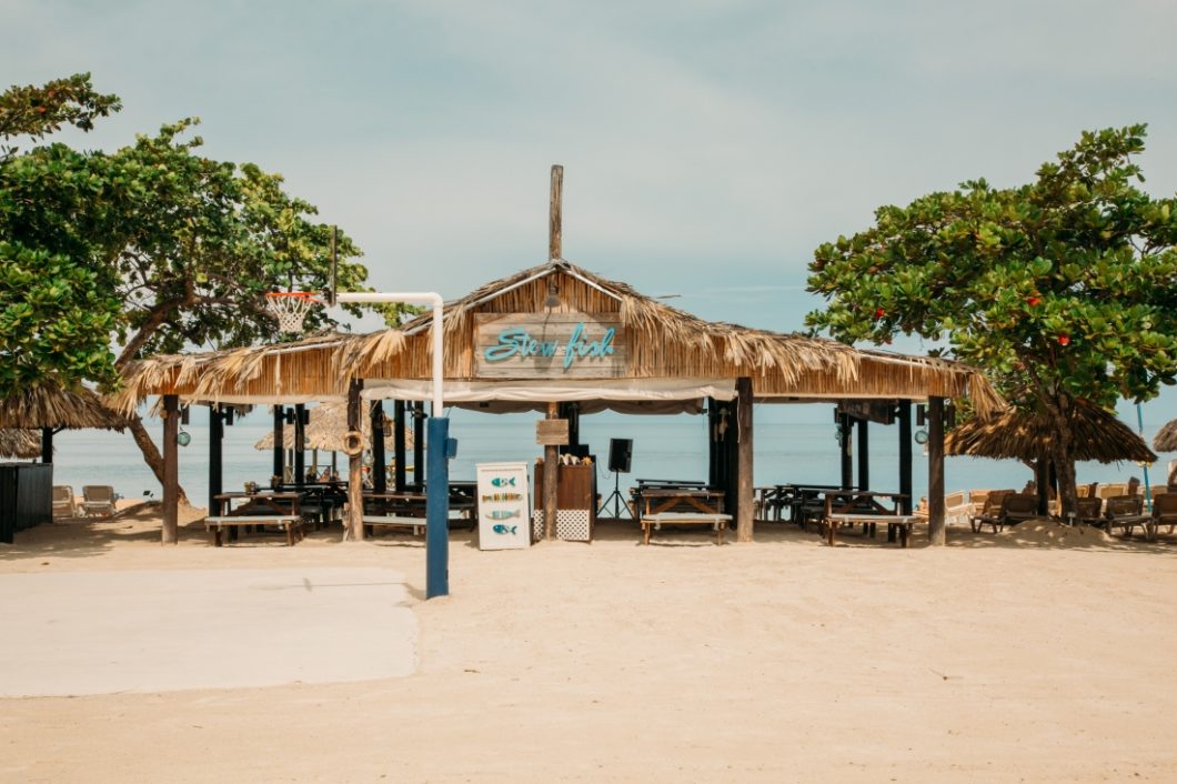 Stewfish restaurant with thatched roof and wooden picnic tables, set beside a sandy basketball court