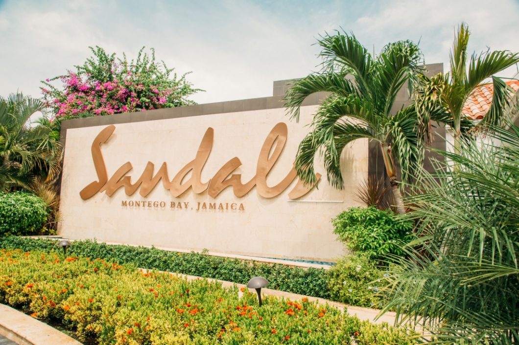 The entrance to Sandals Montego Bay, featuring a large sign that says "Sandals Montego Bay, Jamaica" with palm trees and flowers on each side