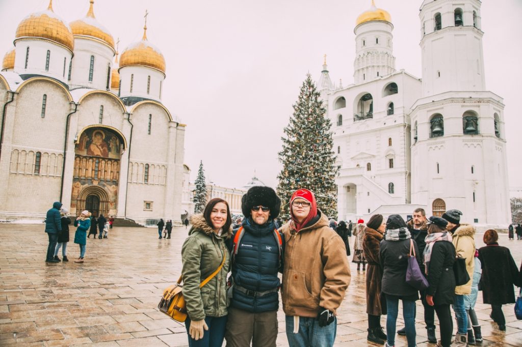 Three people pose in front of two Russian cathedrals during the winter. The square is filled with people, and a large Christmas tree stands in the background.