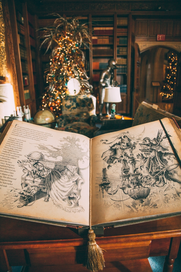 A large storybook is opened, depicting original drawings from The Nutcracker Christmas story. In the background is an elaborate private library with walls of bookshelves, statues, and art. Large Christmas trees stand decorated and lit throughout the room.