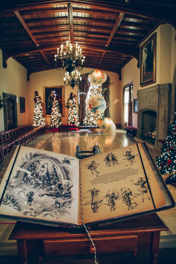 A large storybook is open to a scene in the Nutcracker. The room behind it is vast with a large, open space in the center of the room, meant to symbolize and ice skating rink. The back wall is lined with decorated Christmas trees, while another wall is lined with wooden chairs.