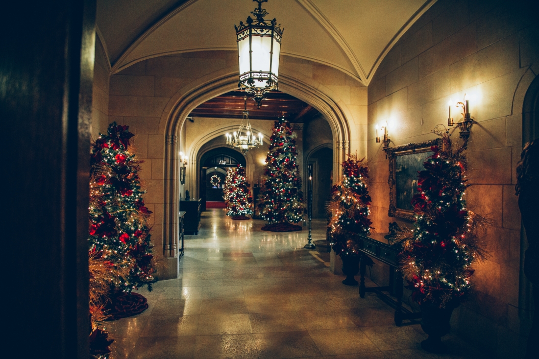 A long hallway with vaulted ceilings and arched doorways, lined with lit and decorated Christmas trees. Antique chandeliers and sconces cast dim, warm light on the walls and floors.