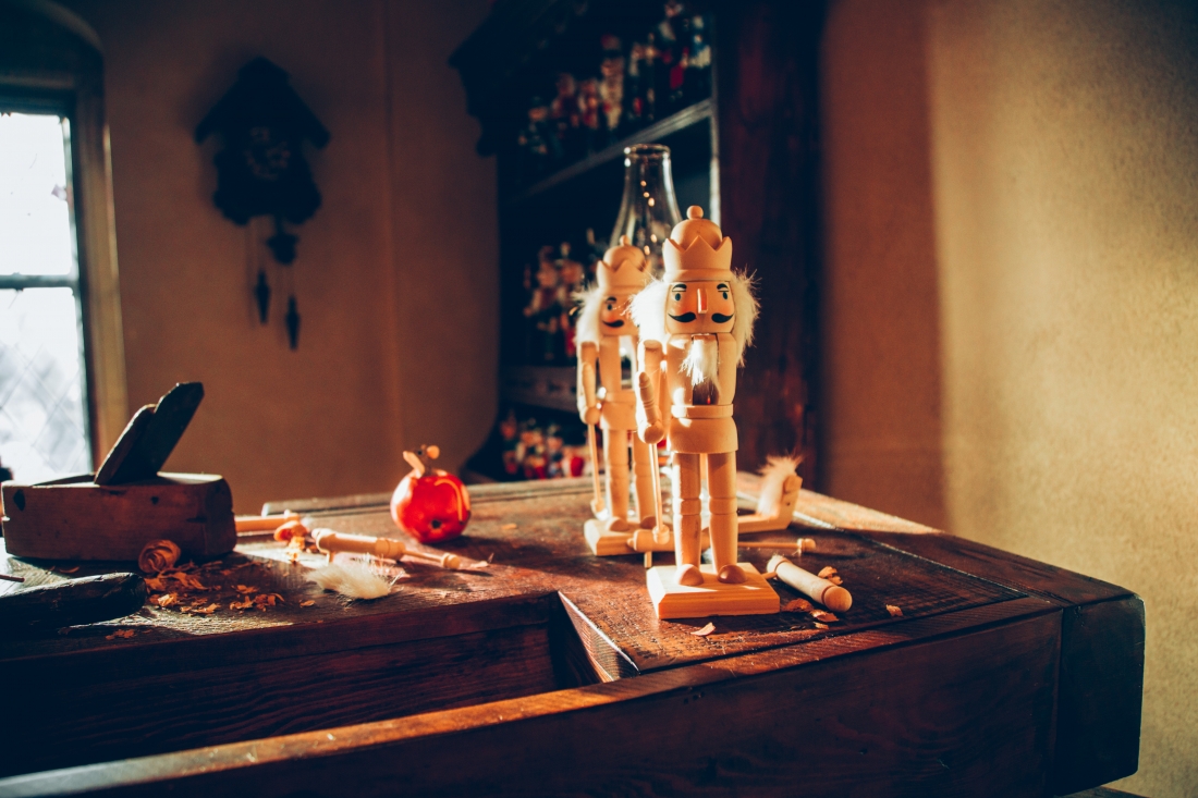 Two small Nutcracker dolls sit on a table next to wooden doll pieces and wood-carving tools.