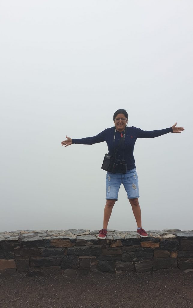 A woman stands on a short brick wall, posing with her arms held out wide in front of thick fog that's obscuring the Haleakala Volcano in Maui 