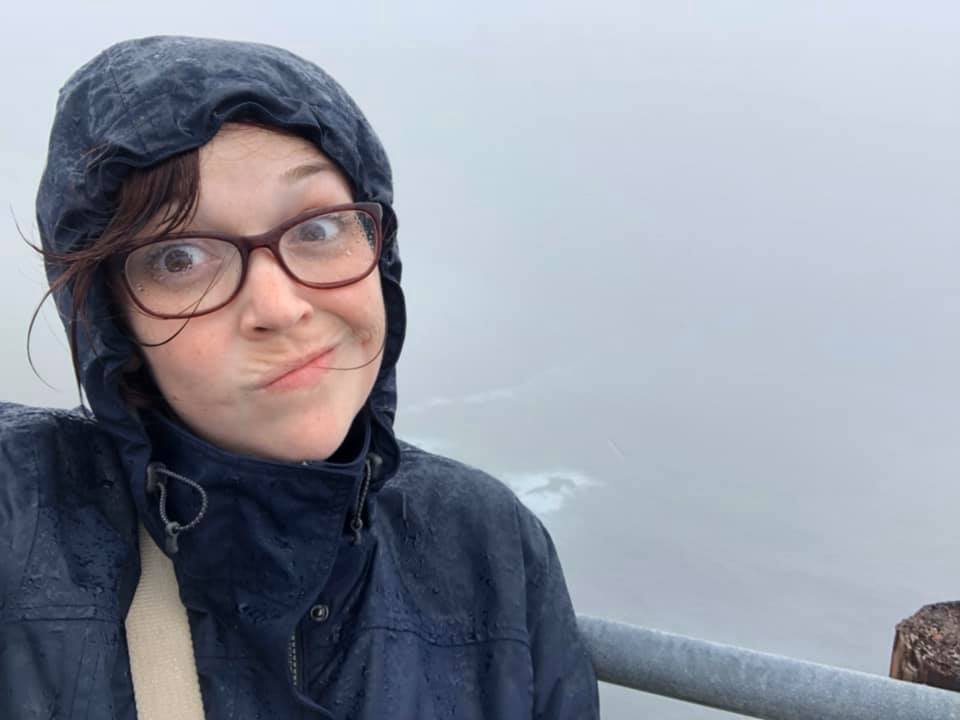 A woman poses in front of the Ring of Kerry in Ireland on a rainy day, where the views of the coastline are obscured by fog and rain clouds.