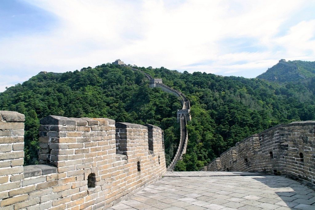 A professional photo of a section of the Great Wall of China that extends upwards through the forest. The wall walkway is empty and the sky is sunny with light clouds.