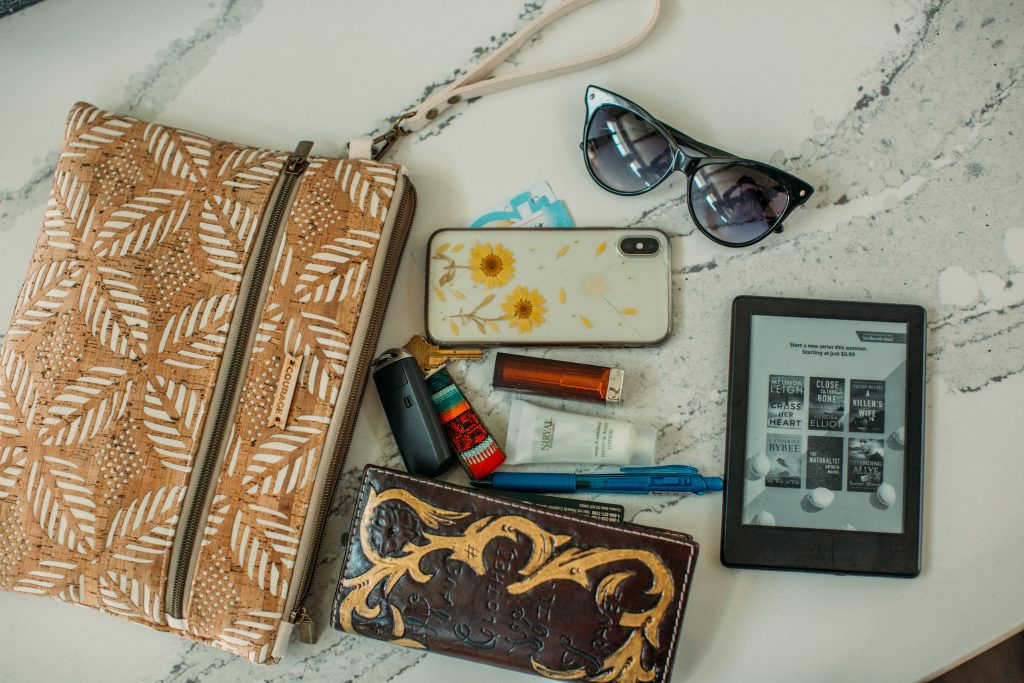 The contents of the INVENTOR Carry Courage clutch bag, showing everything that can fit inside -- a phone, wallet, sunglasses, a Kindle e-reader, and various other purse contents.