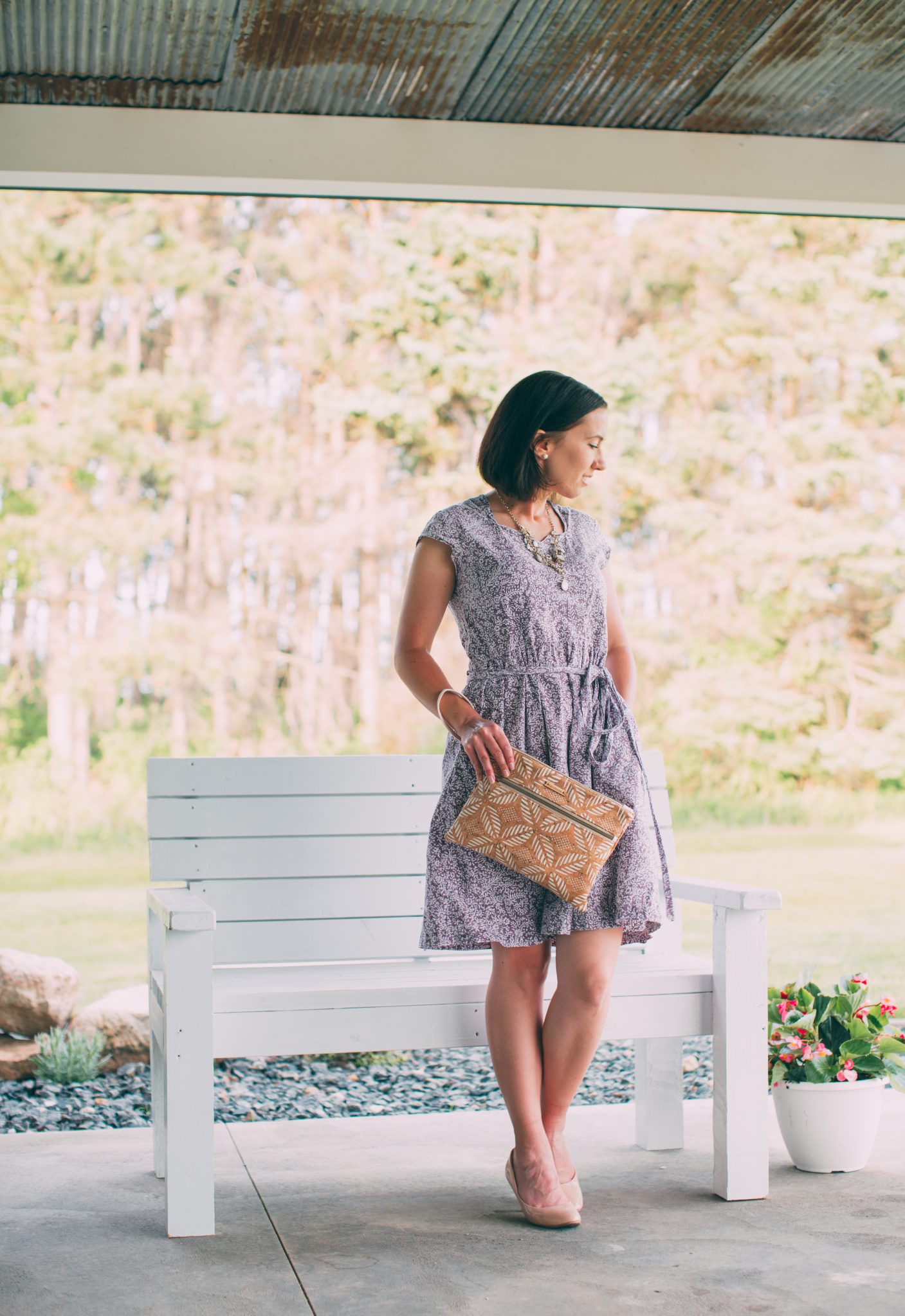 A woman poses outside on a covered patio by a white bench. She's wearing a dusty blue patterned dress and holding the INVENTOR tablet clutch from Carry Courage.