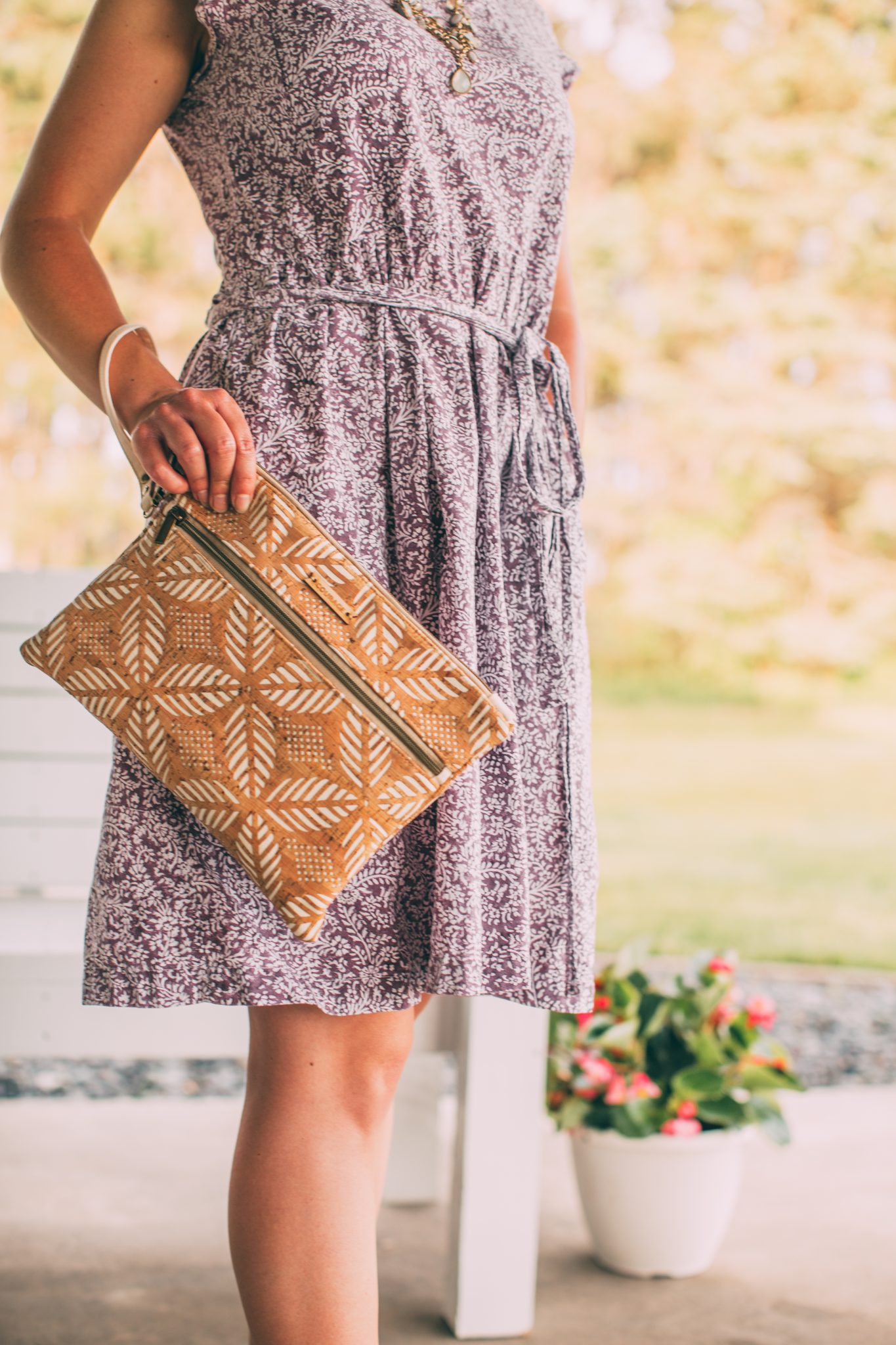 A woman poses outdoors, holding a patterned tablet bag with a thin wristlet strap. The bag has a light brown and tan leaf pattern.