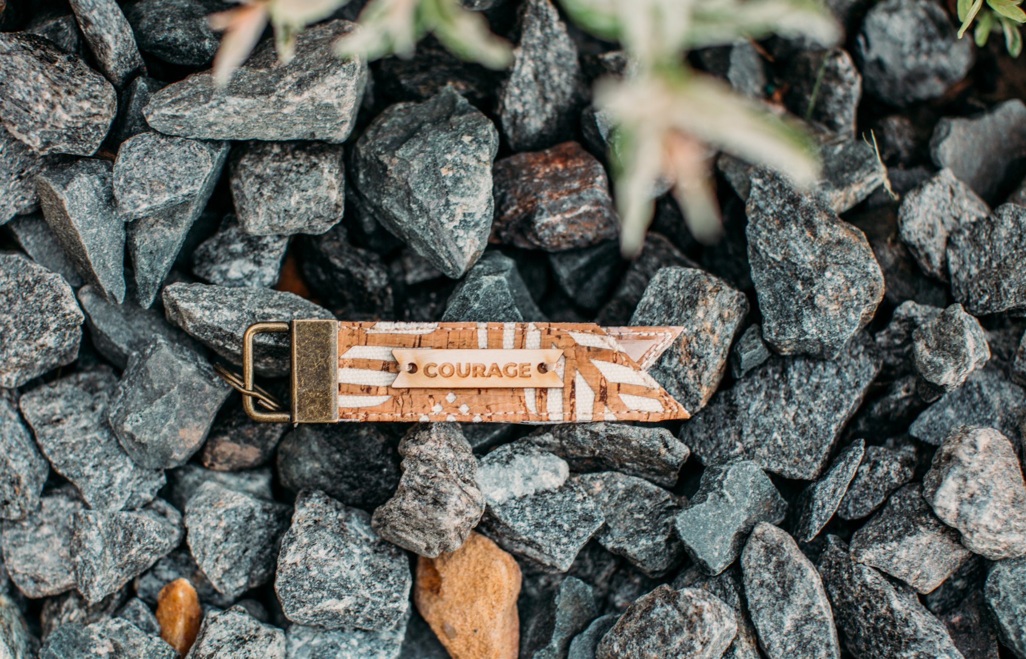 A small keychain luggage tag from Carry Courage sits on a bed of dark rocks, photographed from above.