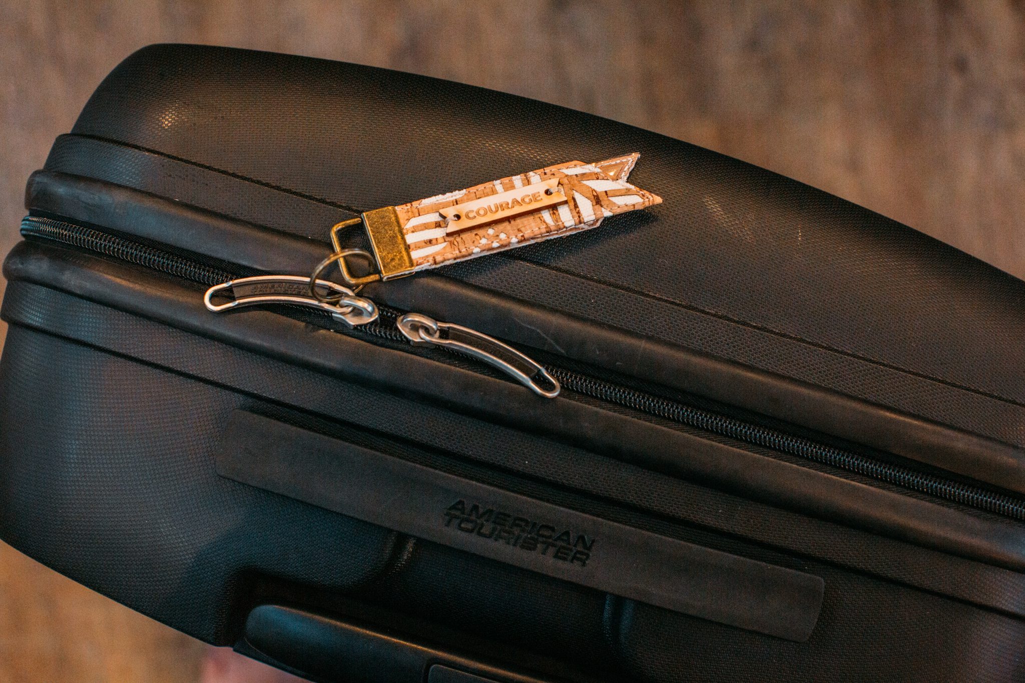 A small luggage tag with a badge that says "courage", attached to the zipper on a black hard-shell American Tourister suitcase.