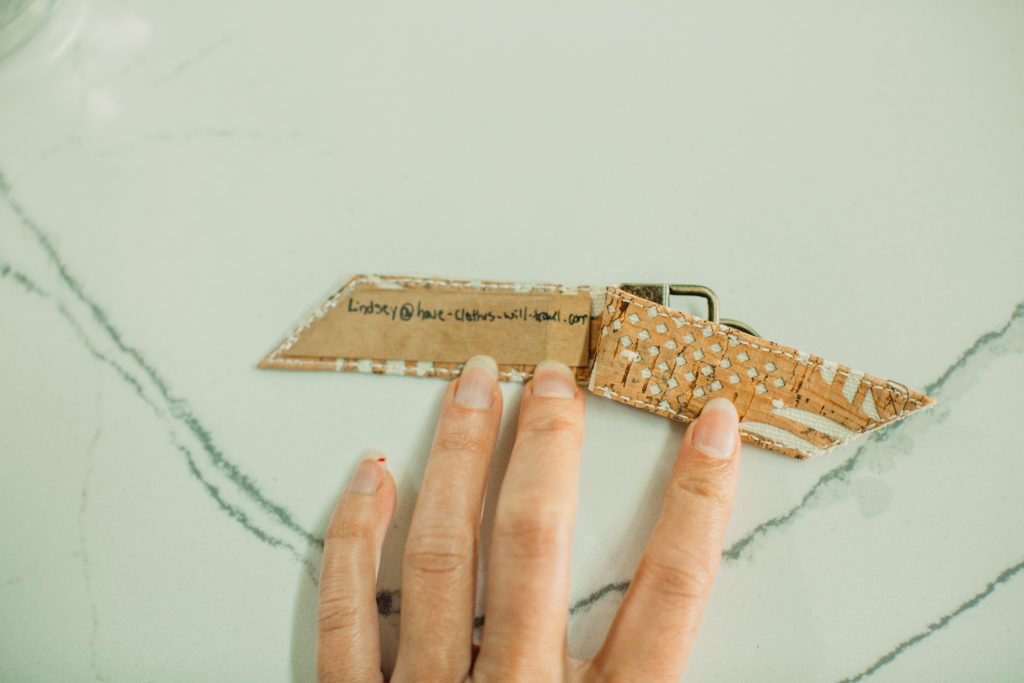 A person holds the Carry Courage luggage tag open against a marble countertop, showing the inside flap where an email address is hand-written.