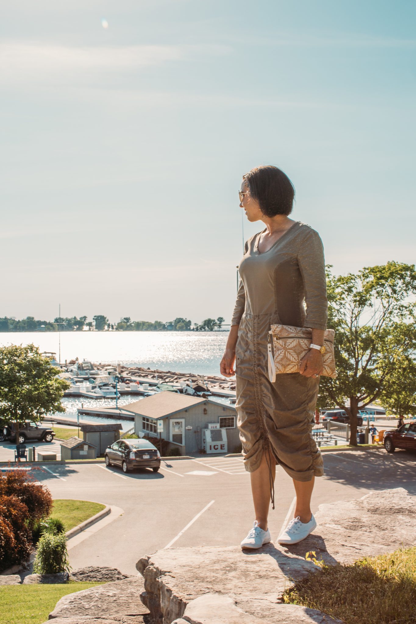 A woman poses in front of a marina in Egg Harbor, WI on a sunny day. She's wearing a brown dress and carrying a Carry Courage clutch bag.