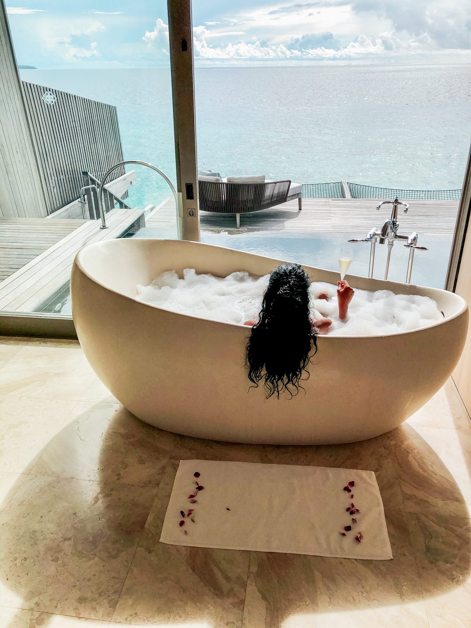 A woman with long dark hair relaxes in a soaking tub with bubbles and holds a glass of champagne. Shee looks out over a private pool and the ocean at The St. Regis Maldives Vommuli Resort in Dhaalu Atoll, Maldives 