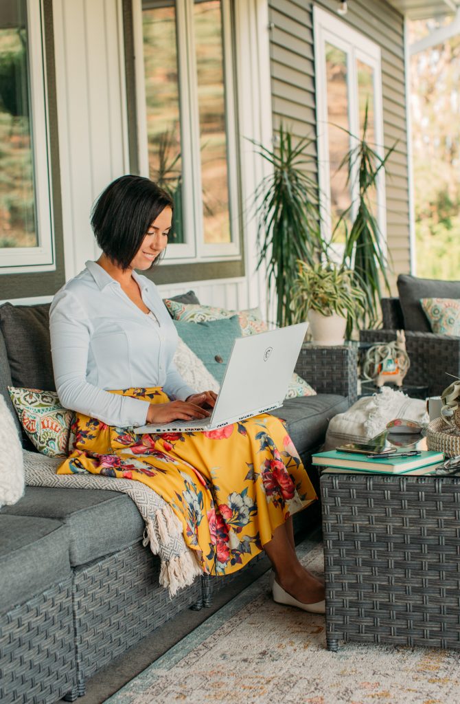 A woman sits on an outdoor couch, with a throw blanket over her lap as she works on her computer in a cozy backyard patio space.