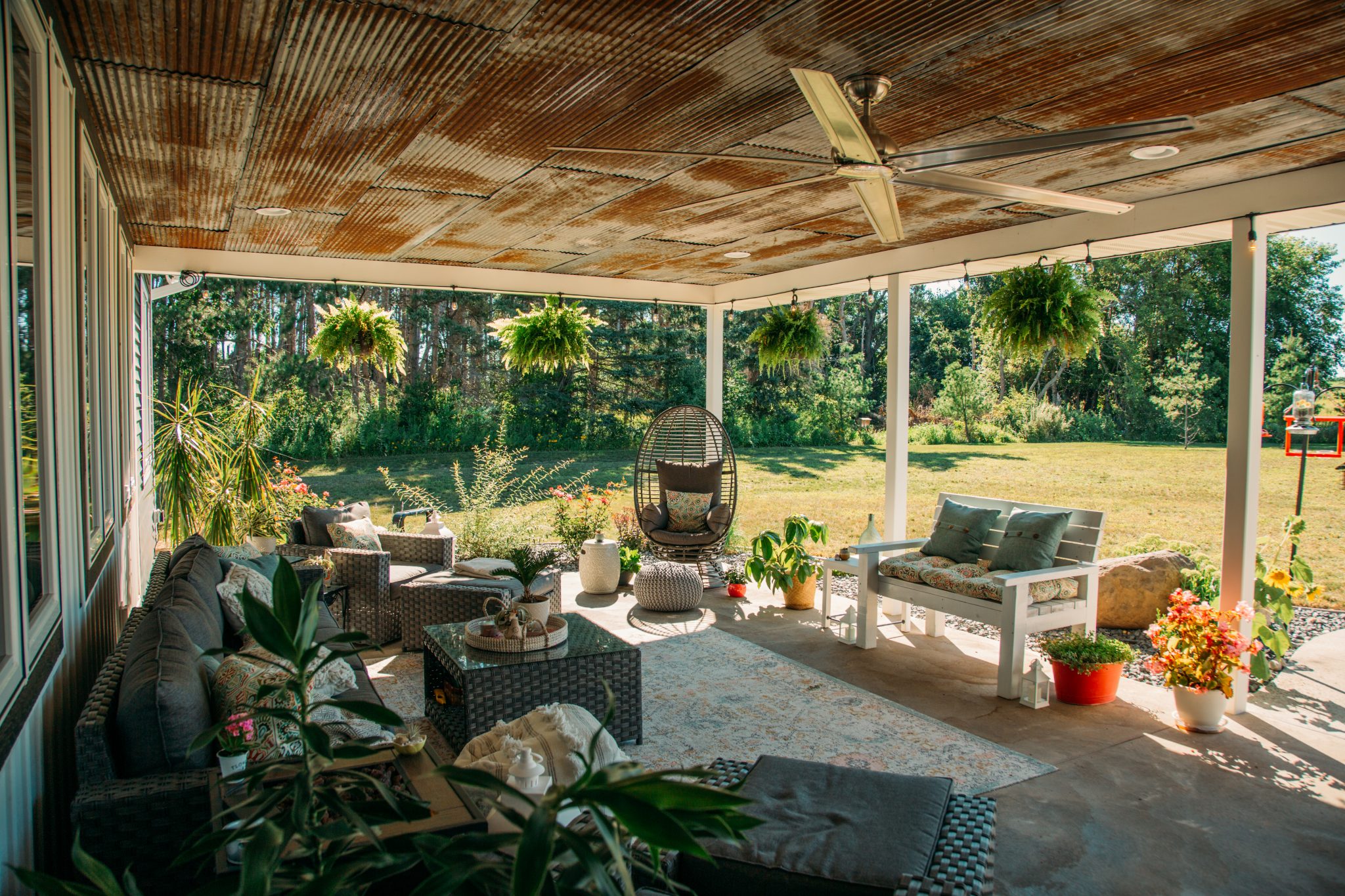 A cozy outdoor patio space on a bright summer day, with a large seating area, hanging planters around the porch and planters of flowers around all of the seating.