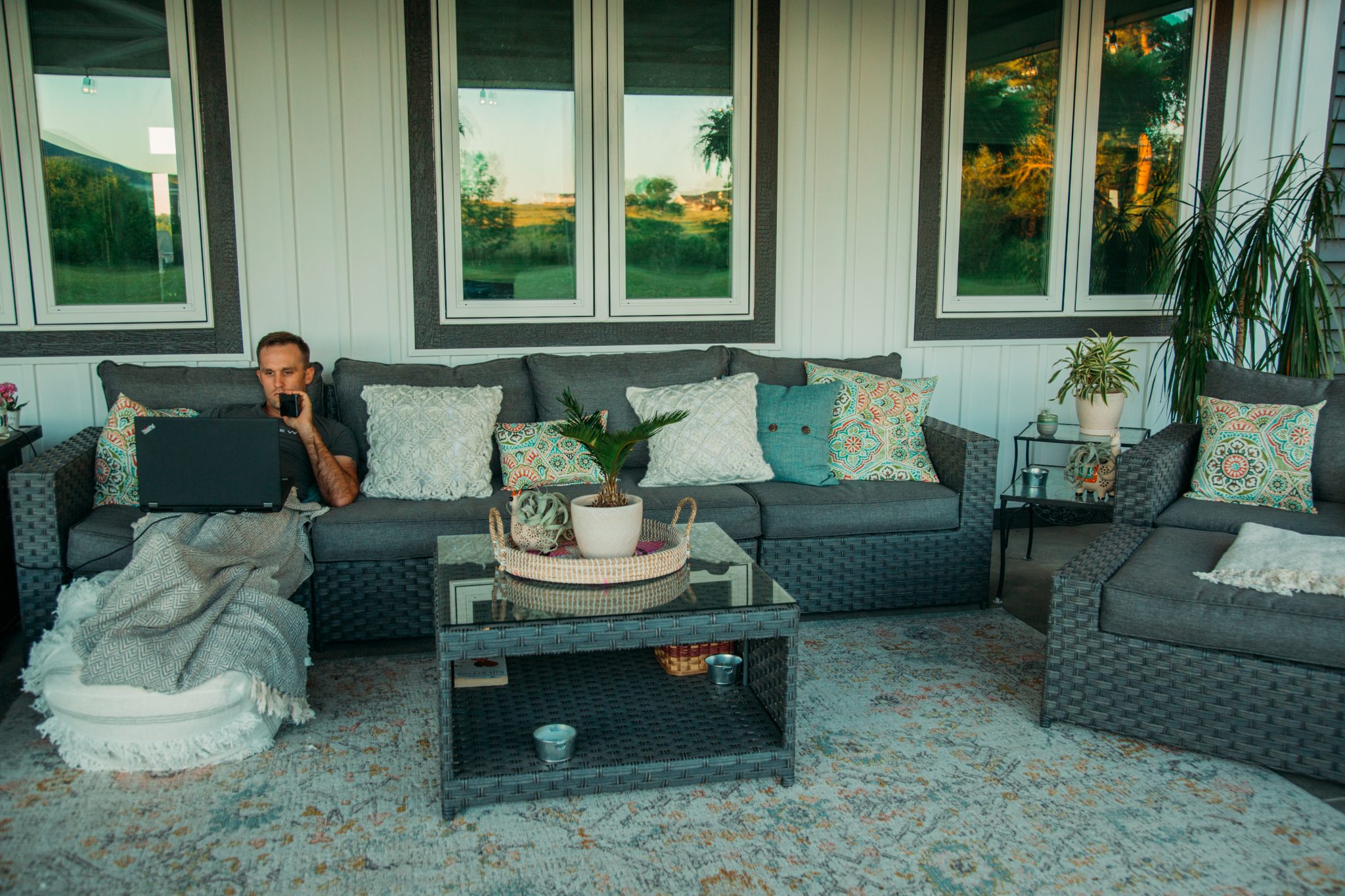 A man sits on an outdoor couch, working on a laptop. He rests his feet on an outdoor ottoman pouf and a throw blanket.