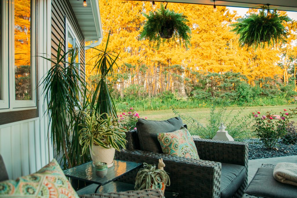 A view from the cozy backyard patio living space, decorated with comfy outdoor furniture, pillows, and plants, overlooking yellow and green trees and landscaping.