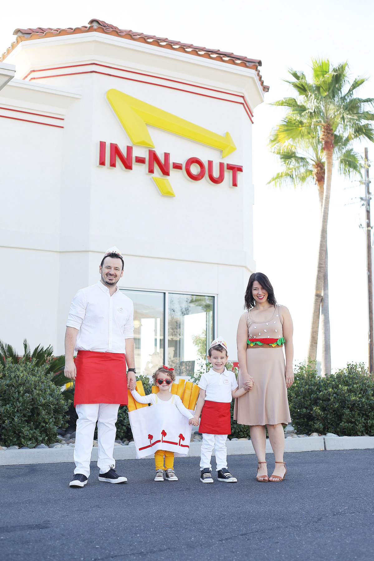 A family of four - mom, dad, and two young children- stand outside of an In-N-Out restaurant, dressed in DIY Halloween Costumes. The dad and little boy are dressed as fast food workers, the mom wears a dress that looks like a burger, and the little girl is dressed in a French fries costume.