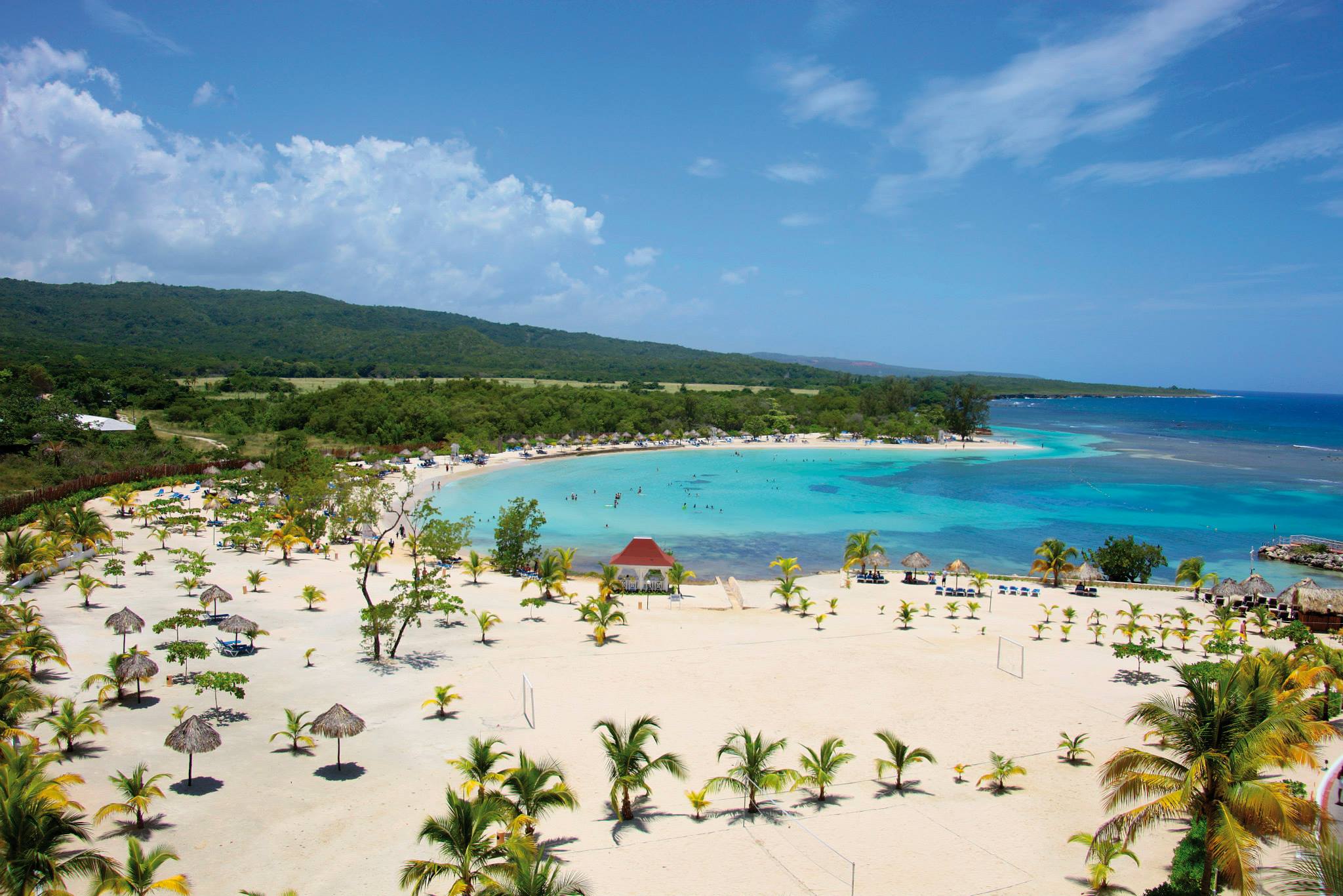 An aerial view of the large private beach area at Bahia Grand Principe in Jamaica. The beach features plenty of lounge areas, volleyball nets and recreational areas, and a clear blue inlet for swimming.