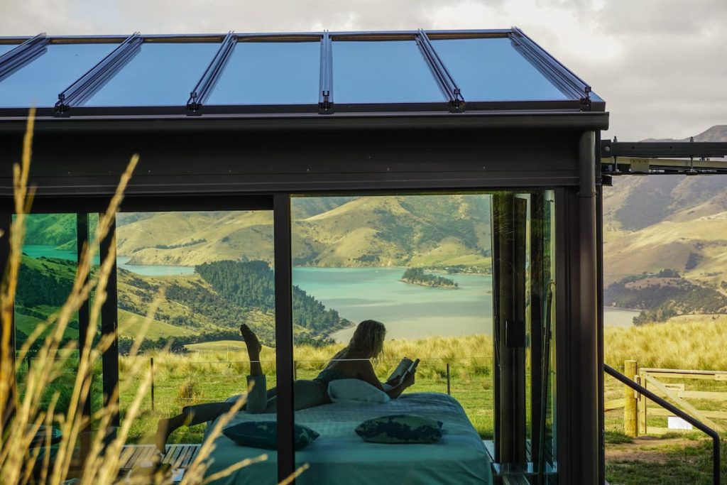 Pōhue PurePod in New Zealand, a glass pod cabin, with a woman reading on bed and views of a lake and mountains in the background