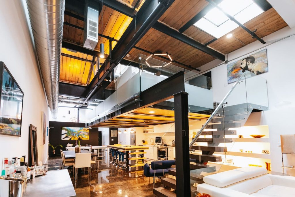 A modern loft space in Toronto features black painted exposed steel beams, natural wood ceilings, and white walls. The room has a kitchen and dining area under a raised sleeping loft sectioned off with a glass railing.