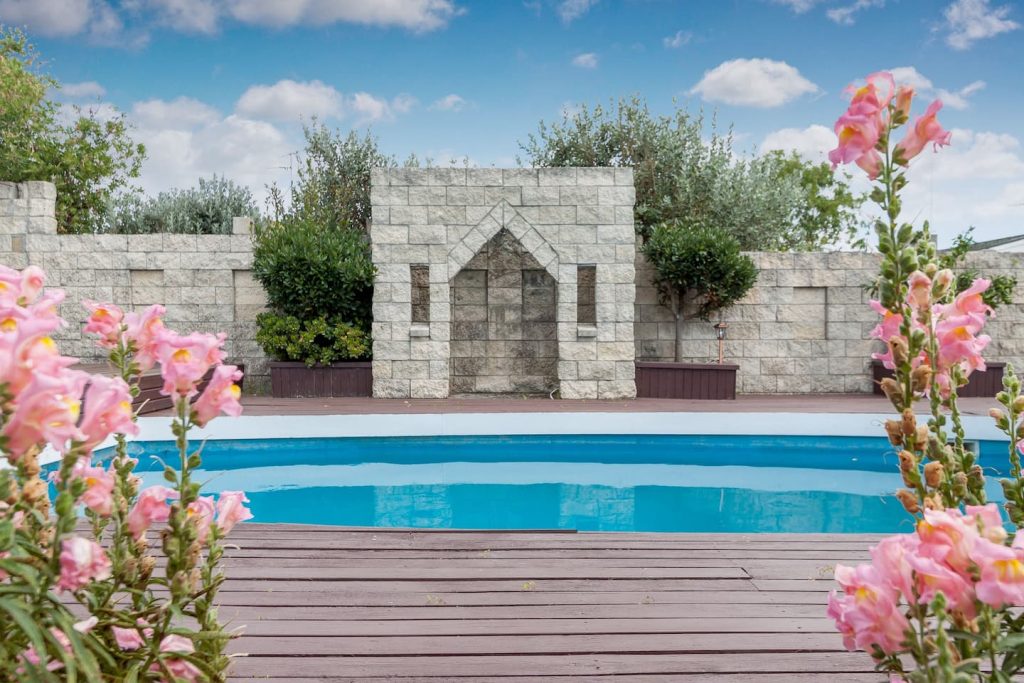 A pool set in a flower garden with brick, castle-like structures in the back