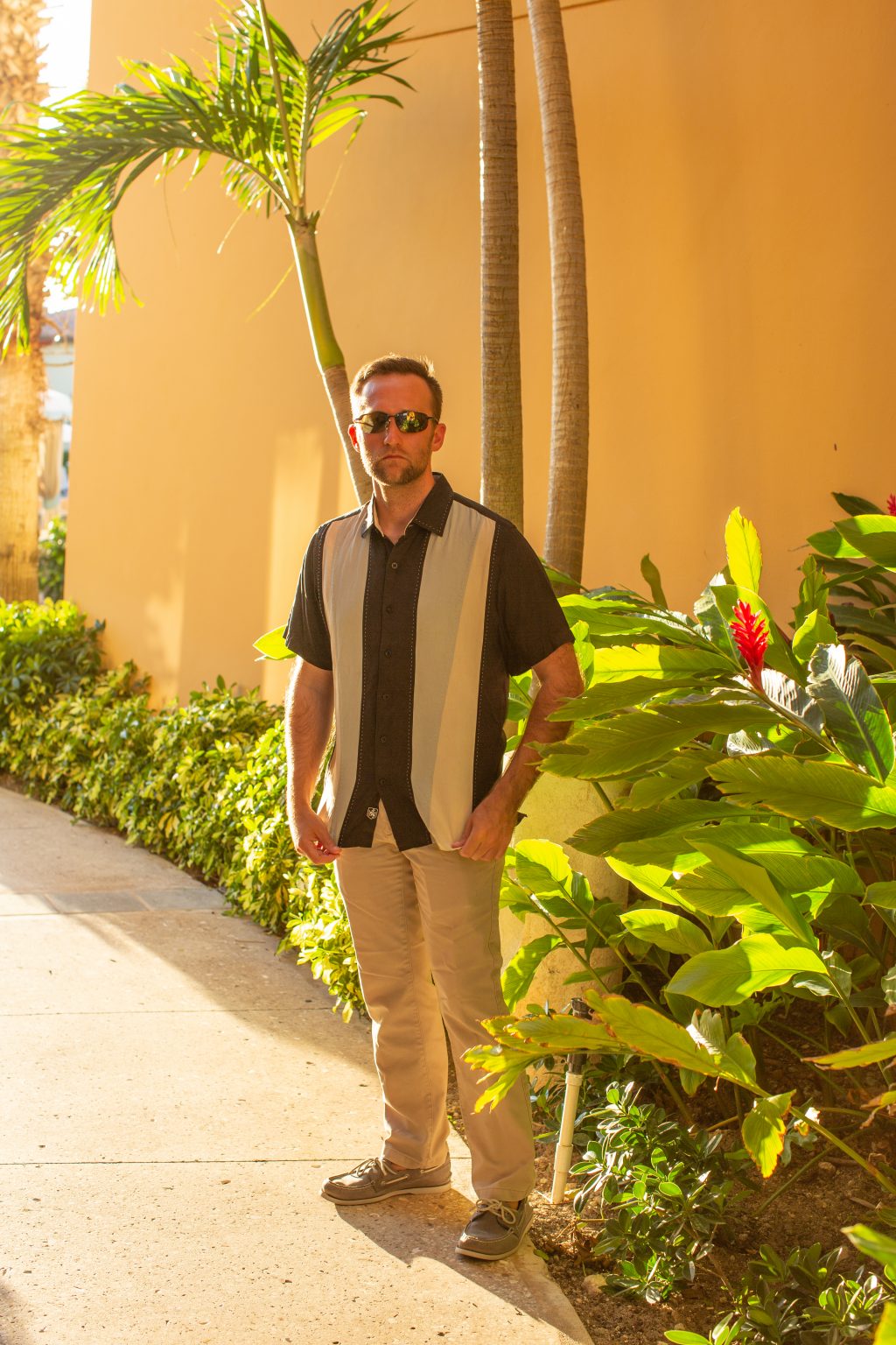 Zac wearing a bowling shirt, chino travel pants, and boat shoes standing next to a palm tree and other tropical plants