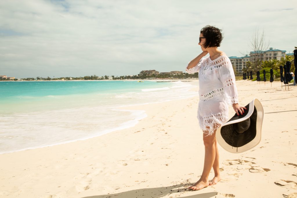 A woman wearing a white cover up holding a hat on the beach.