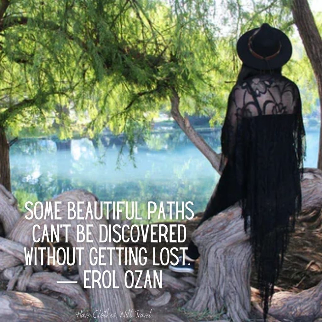 A woman wearing all black overlooks a lake from a tree-shaded viewpoint. Text across the image reads, "Some beautiful paths can't be discovered without getting lost. - Erol Ozan"