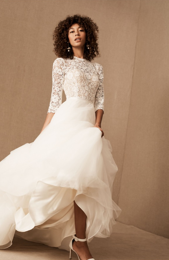 A model wears a full white ballgown wedding dress. The tulle skirt is full and puffy, and the lace bodice has three-quarter length sleeves and an illusion sweetheart neckline.