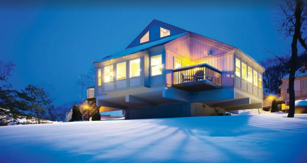 A cozy one-story cabin at dusk. The cabin is illuminated from the inside and features walls of windows and a small outdoor patio with chairs. 