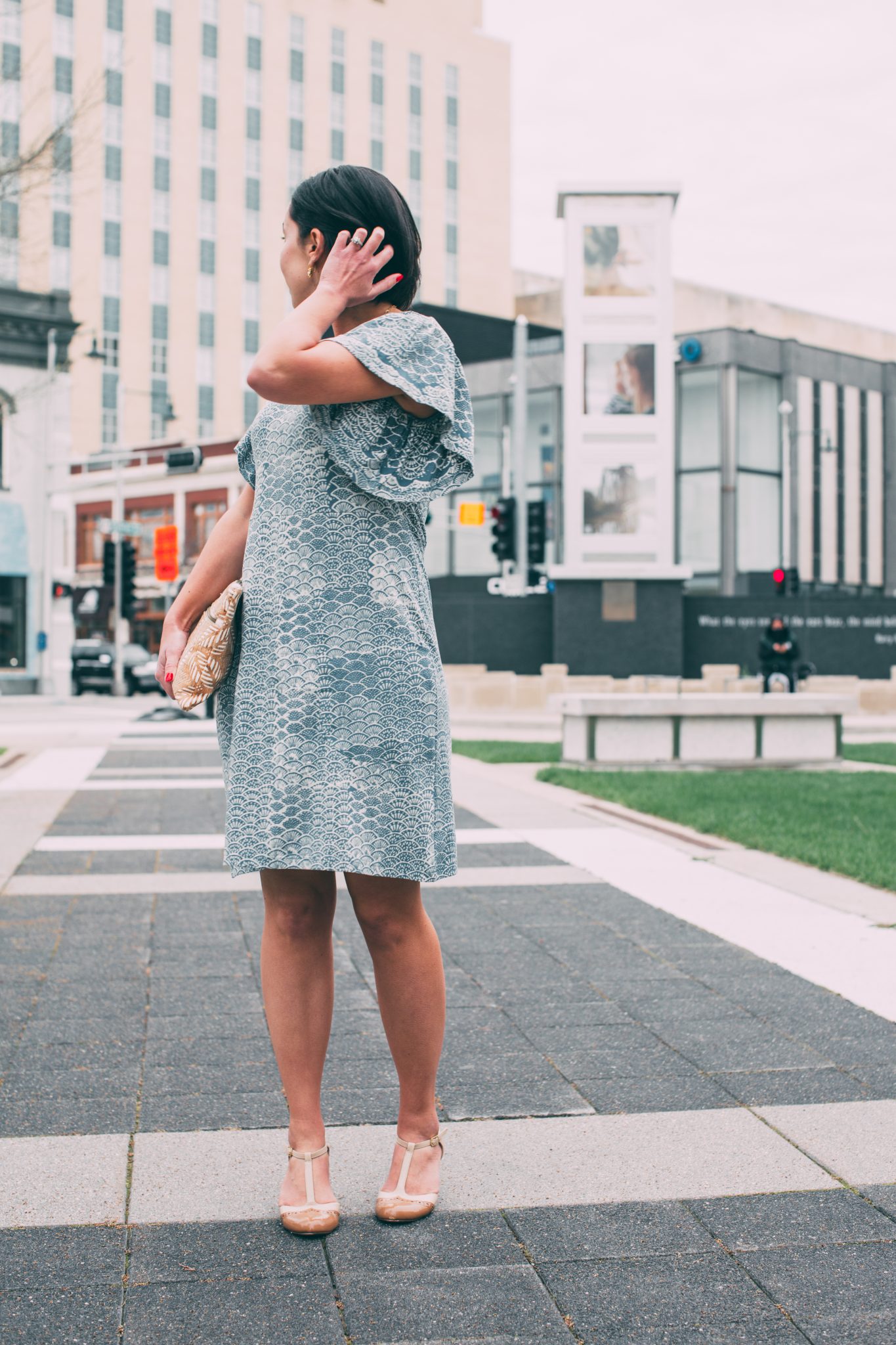 A woman stands on a city sidewalk, looking over her shoulder behind her. She's wearing a blue patterned dress dress and high heels.