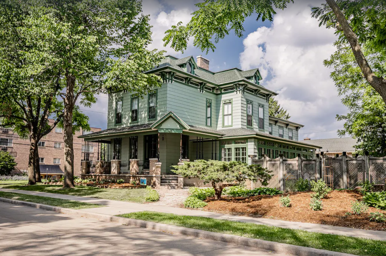 An exterior shot of a Victorian style home, shrouded by trees against a cloudy blue sky.