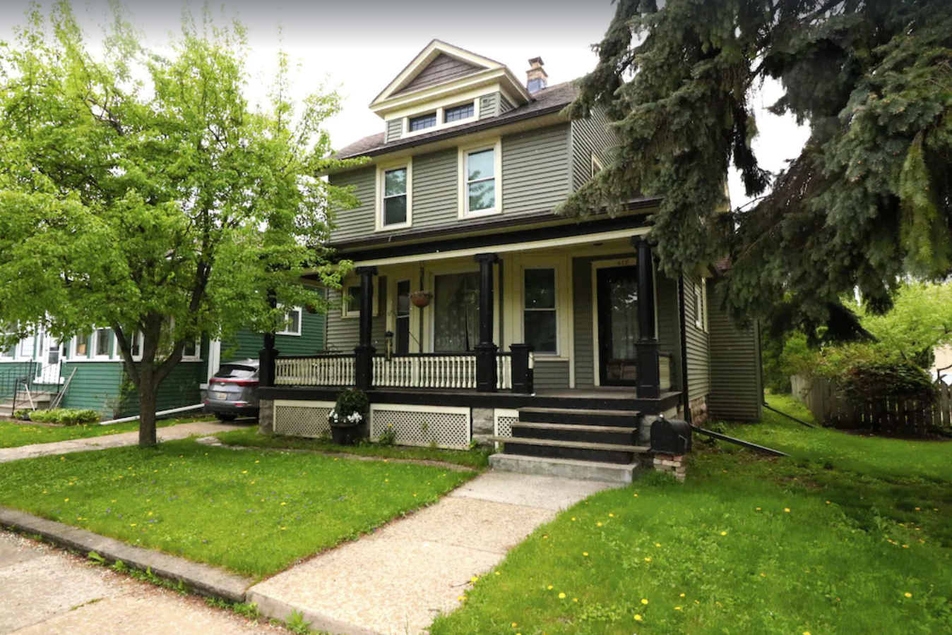 An exterior image of a modest two-story 1900s home with olive green siding and white trim. The home has a small porch and small front lawn with large trees.