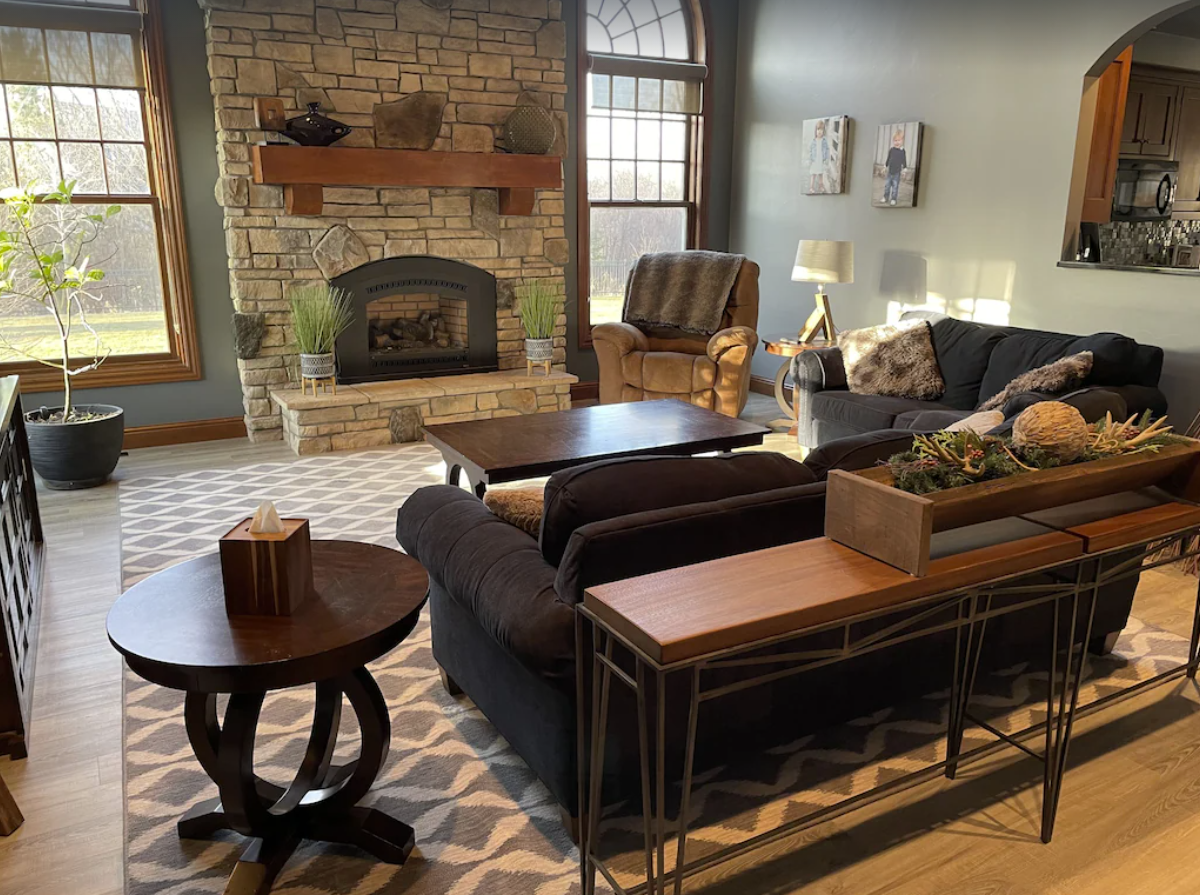 A modern living room with two black fabric couches arranged in front of a stone fireplace. The home is decorated with modern style decor, end tables, and plants throughout.