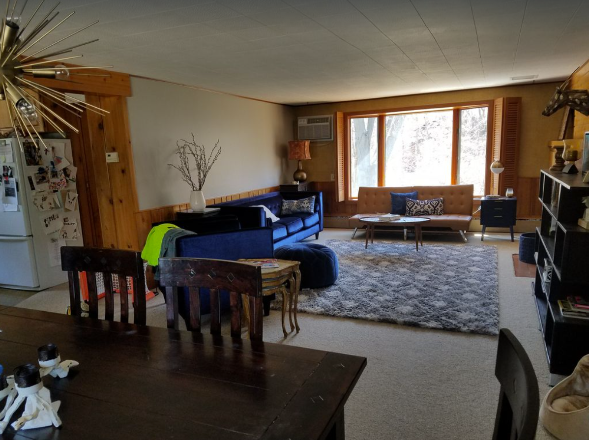 An interior image of a casual living room setting with mismatched couches, a coffee table, and dining room table. A bay window lets sunshine into the room.