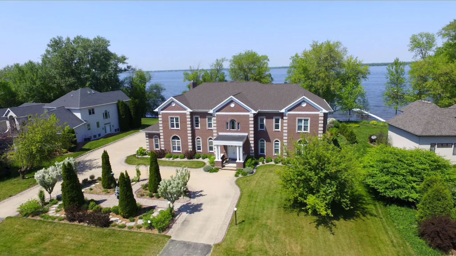 A stunning aerial exterior image of a large two-story, five bedroom home on a lakefront property. The home has a brown exterior and dark asphalt roof, a long driveway and front courtyard with trees and landscaping. 