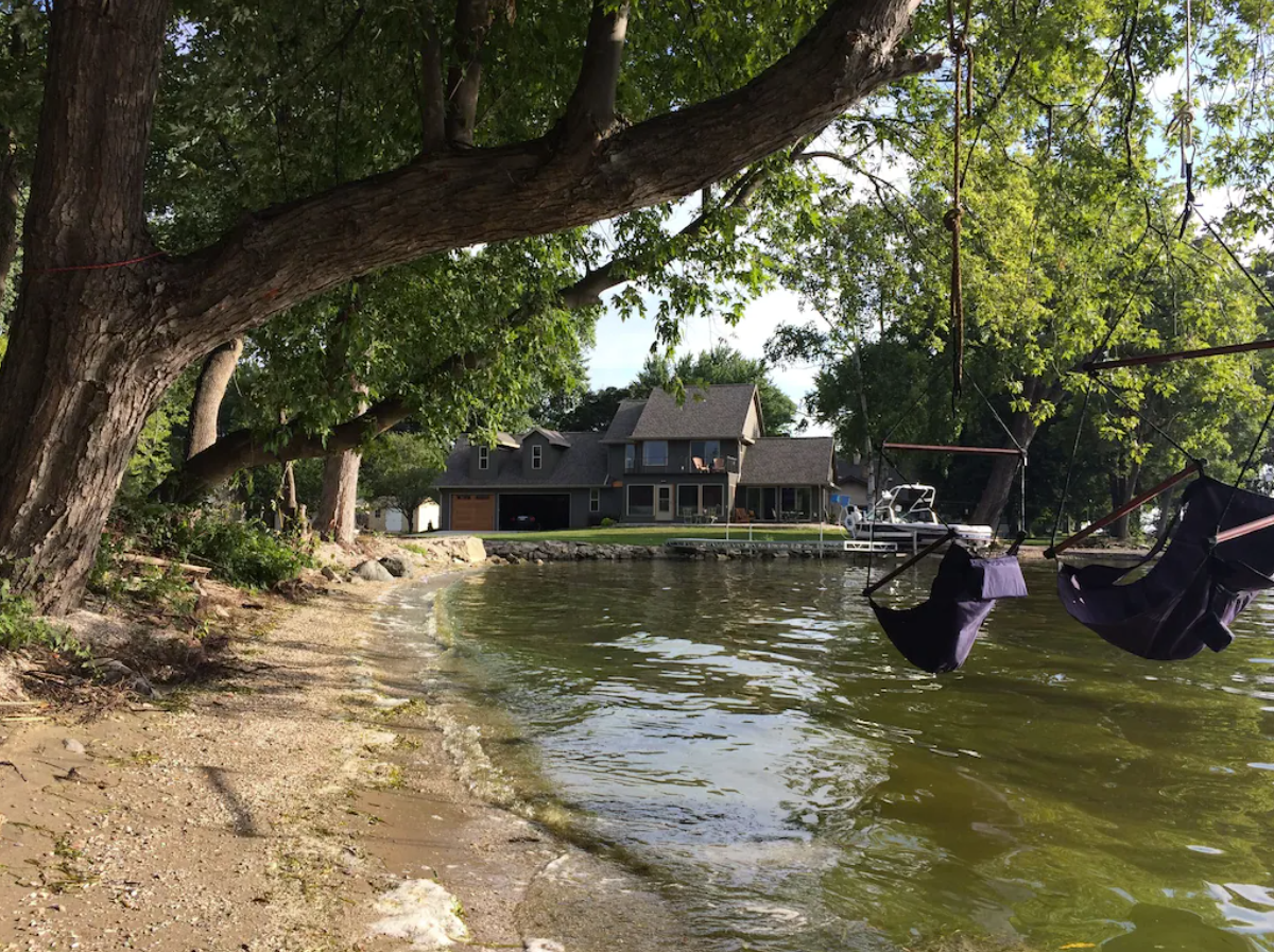 An exterior image of a lakefront property. The house in the background is a quaint two-story home with a brown and gray exterior. It sits right on a small lake with a wooded shoreline. In the foreground of the image are two swings that hang from tree branches out over the water.