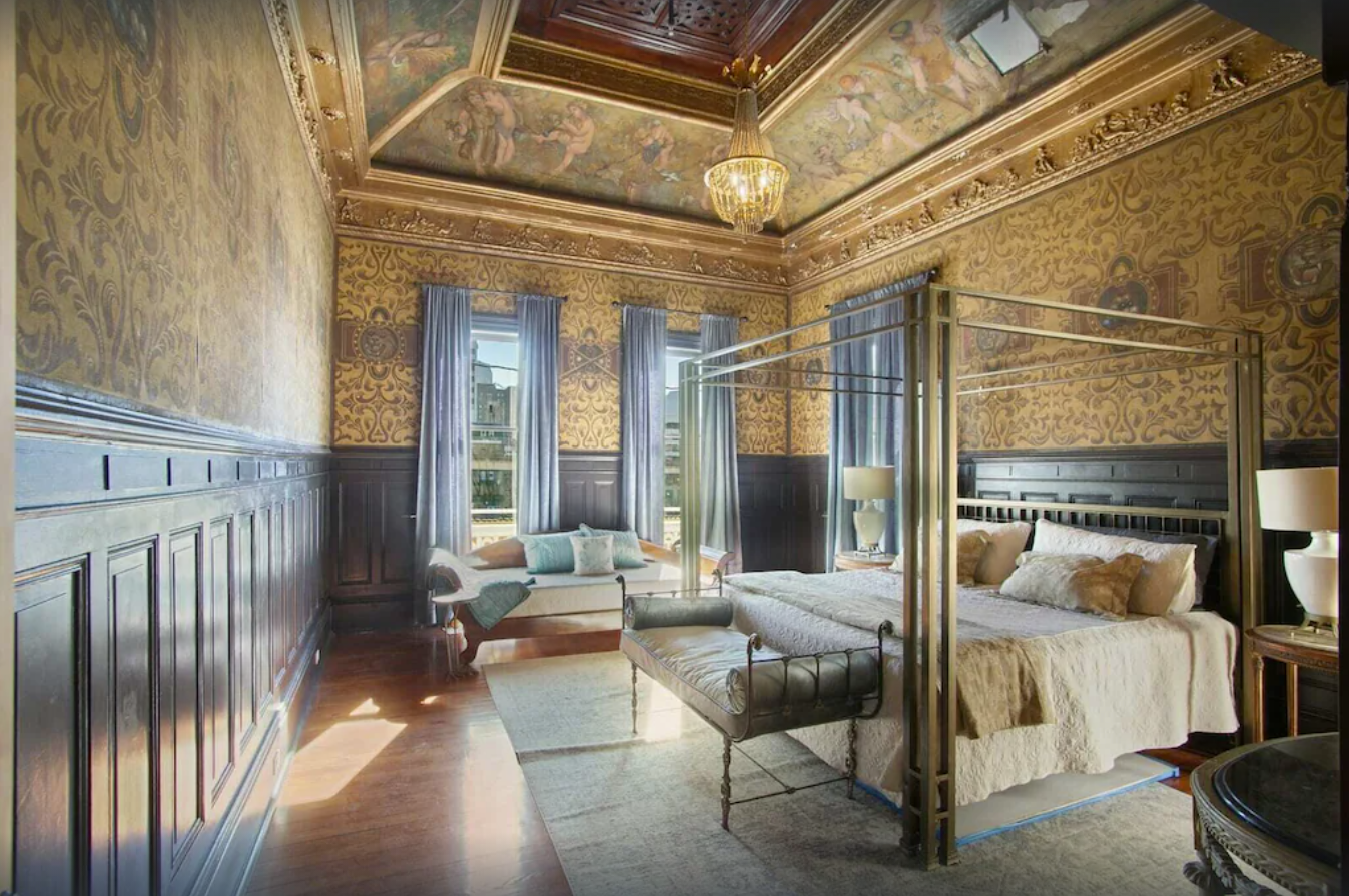 An interior image of large master bedroom with large canopy bed and vaulted ceilings. The walls are covered in fleur de leis wallpaper, the ceiling has stunning gold crown molding and mural artwork around the top of the room.