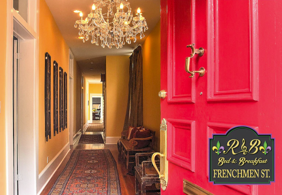 An image shows the entrance hallway of a New Orleans rental home. The red front door opens to a long hallway with a crystal chandelier and yellow walls lined with artwork and benches.