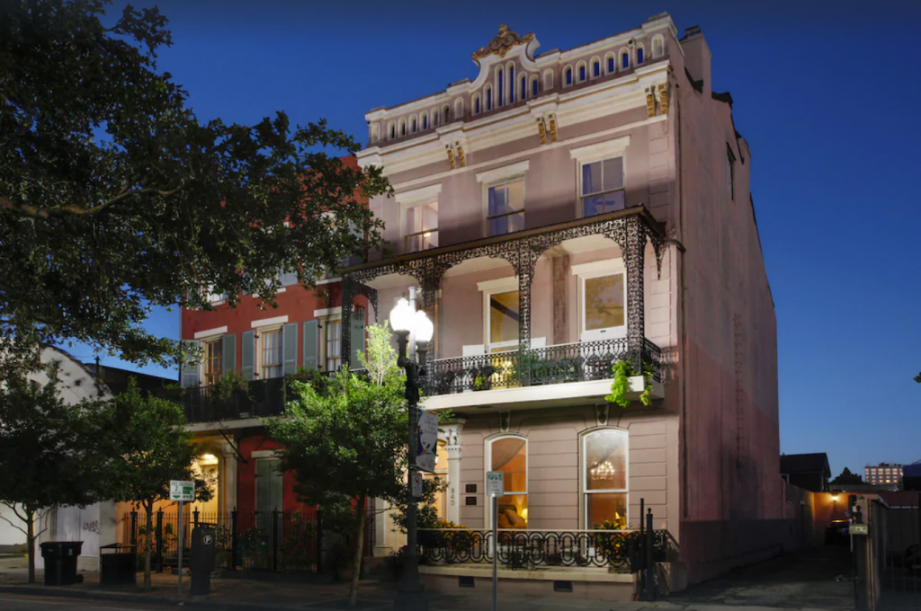 An exterior shot of a 4-story Greek mansion in New Orleans at nighttime. Each level has an outdoor balcony and large windows. The outer façade is a crème white stone.