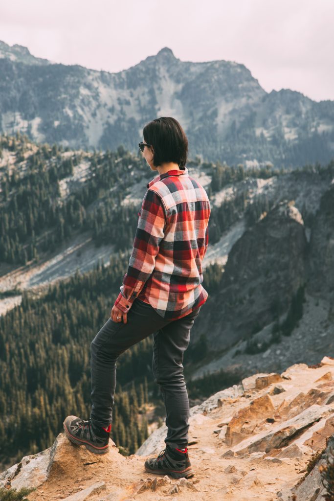 Lindsey standing on a rock looking out over a mountainous landscape, she is wearing a red plaid shirt, grey pants, and hiking boos. 