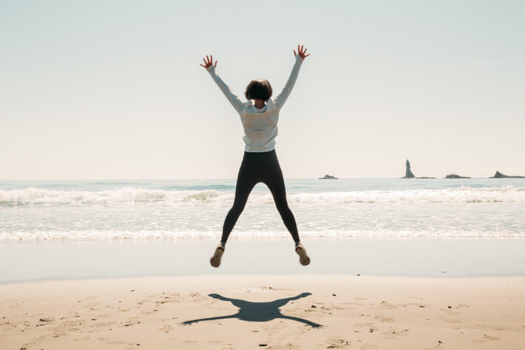 Lindsey wearing black leggings and a tan long sleeve shirt, she is jumping with her hands outstretched on a the beach in La Push, Washington